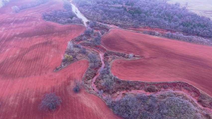 A smooth aerial drone shot flying over rolling red clay farmland with a winding creek cutting through the landscape. Bare winter trees and muted seasonal colors create a rugged rural aesthetic. 