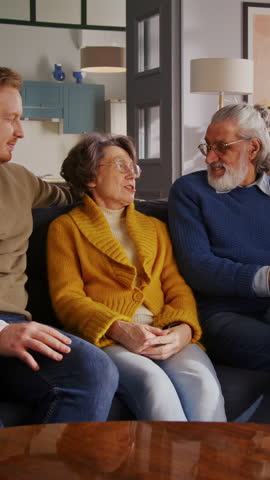Caucasian family calmly resting on pillowed couch and getting amused by active kids running from another room. Cheerful sister and brother jumping over back of sofa for getting attention from adults.