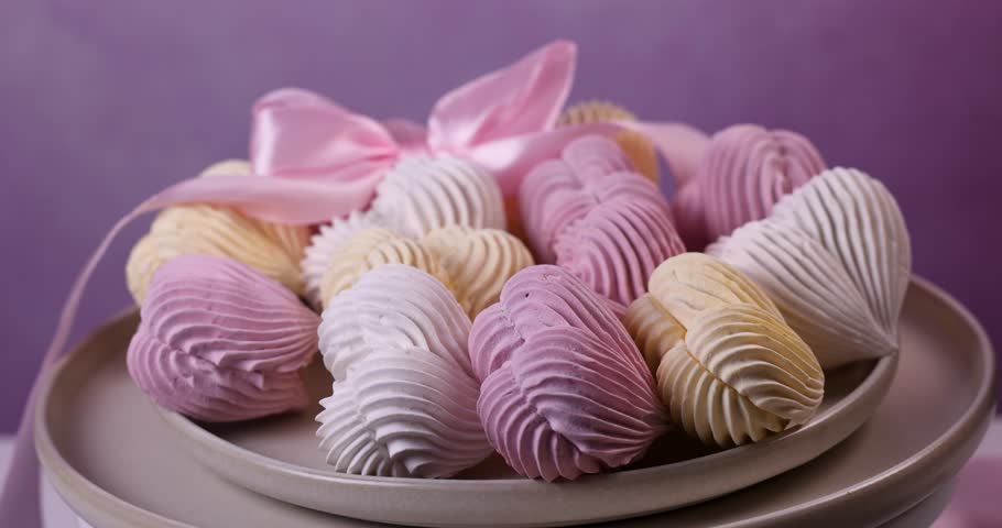 Female hand with pink manicure gently taking a pastel colored, heart shaped meringue from a ceramic plate filled with assorted sweet cookies against a beautiful purple background