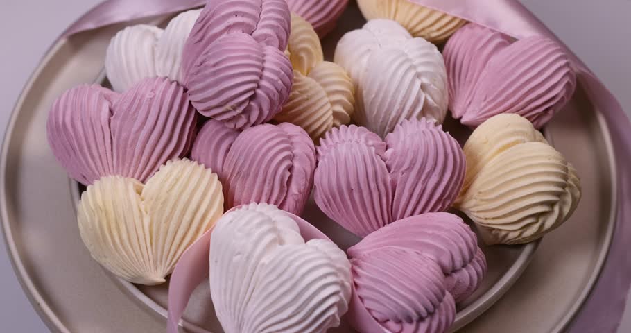 Female hand placing a red love sign among delicate, colorful, heart shaped meringue cookies arranged on a plate with a pink satin ribbon for a romantic valentine