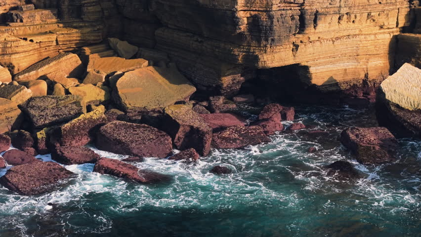 Aerial close-up of vibrant layered rock formations at Cabo Carvoeiro with rugged yellow and red stone blocks meeting swirling turquoise ocean water along the dramatic Portuguese coastline