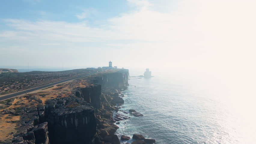 Expansive aerial view of Cabo Carvoeiro cliffs and lighthouse with long coastal road, rugged shoreline, and bright ocean haze creating a dramatic and atmospheric Portuguese seaside landscape