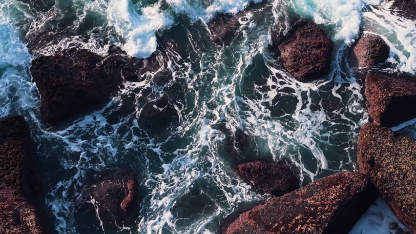 Aerial close-up of powerful turquoise ocean waves swirling and crashing against dark red coastal rocks, creating dramatic white foam patterns along the rugged shore of Cabo Carvoeiro