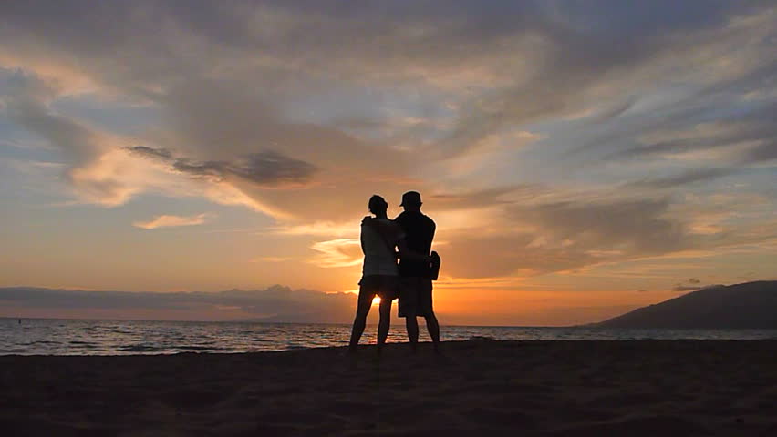 Man and woman stand together, watching sunset on beach in Hawaii.