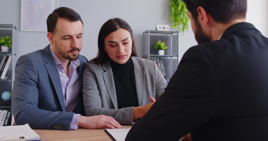 Serious young couple listening to male advisor during consultation in office. Man and woman sitting at desk together and focusing on specialist explaining details of financial or legal matter.