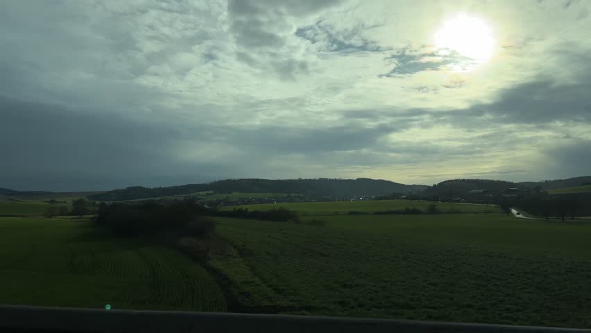Peaceful rural landscape with rolling green fields, distant hills, and a bare tree silhouetted against a cloudy sky on an overcast day.