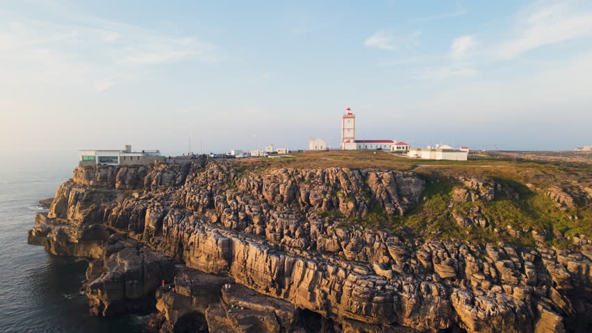 Aerial sunset view of Cabo Carvoeiro lighthouse and rugged coastal cliffs glowing in warm golden light above the Atlantic Ocean, highlighting Portugal’s dramatic shoreline and rocky landscape