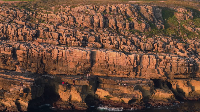Aerial sunset close-up of Cabo Carvoeiro’s rugged stratified cliffs glowing in warm golden light with people standing on rocky ledges above the Atlantic coastline in Portugal