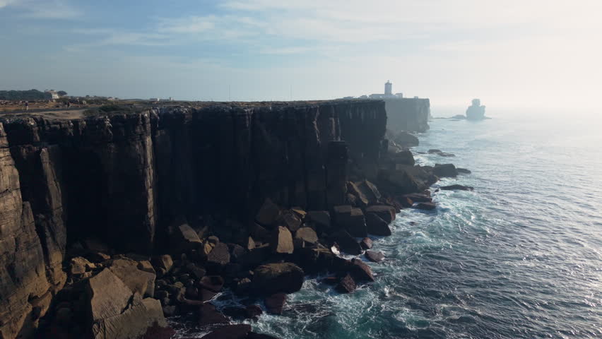 Dramatic aerial view of the towering rocky cliffs of Cabo Carvoeiro rising above the Atlantic Ocean with waves crashing against the rugged shoreline under soft coastal haze on a clear and tranquil day