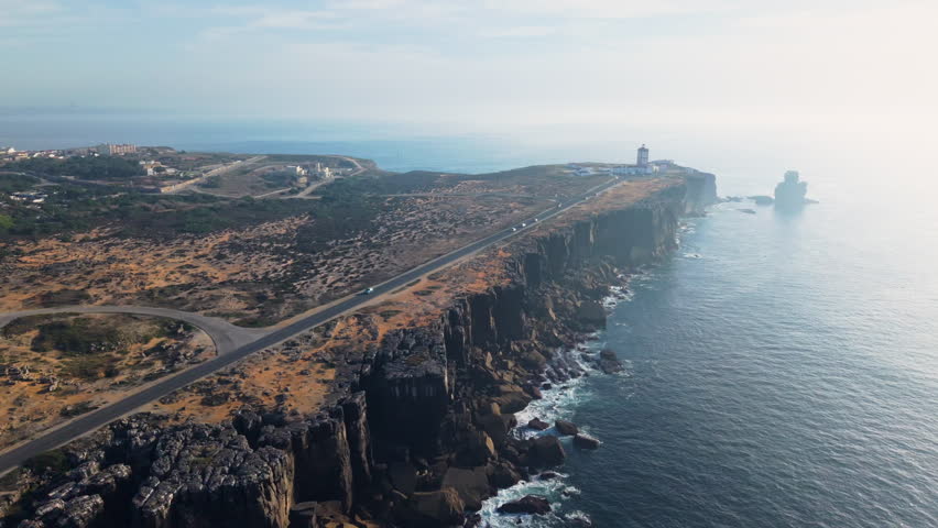 Aerial view of Cabo Carvoeiro cliffs with coastal road, lighthouse, rugged rocky shoreline, and calm Atlantic Ocean under soft morning haze creating a dramatic and expansive coastal landscape