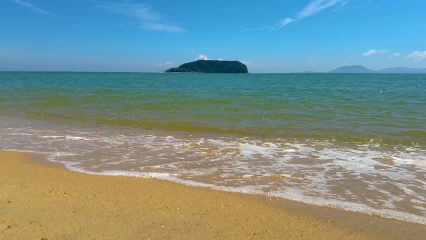 Gentle waves washing onto a sandy beach with a distant island under a clear blue sky.