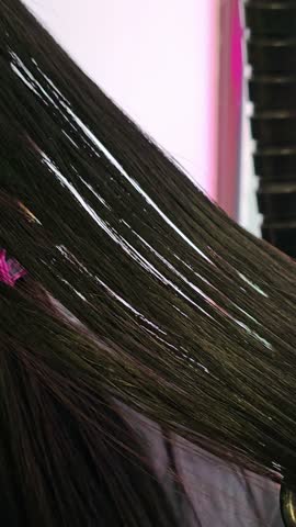 Hair stylist blow-drying and styling a woman's long hair in a beauty salon. Vertical close-up video of stylist using hair dryer and comb to dry and shape dark hair in beauty salon