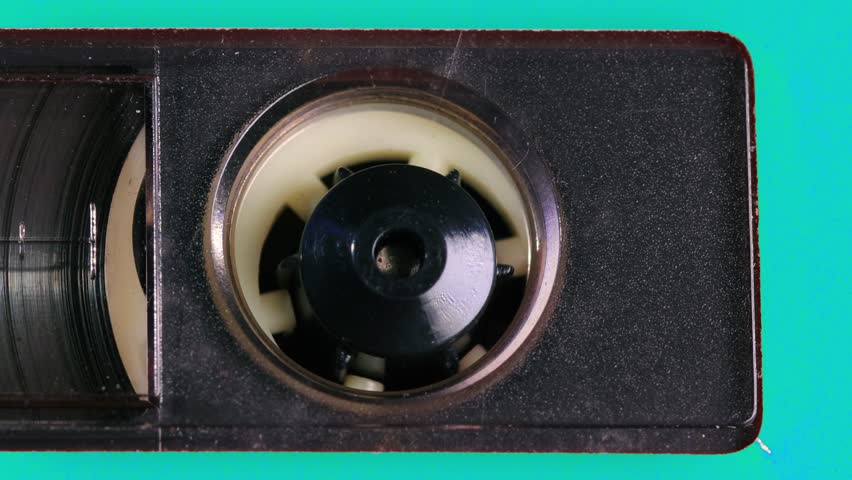 Nostalgic audio cassette tape unwinds behind a transparent window in a turquoise blue plastic cover. Tapes reels, percent marks coming into view as the camera pans left. Extreme Close up
