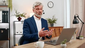 Middle-aged smiling businessman at home office wears headset assisting hotline caller via laptop. Freelancer guy at table gives rapid support ensuring customer satisfaction enhancing service quality - Powered by Shutterstock - Get 15% off with code: PIKWIZARD15
