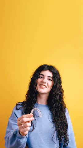 Cheerful young woman with a beautiful smile holding and presenting a transparent dental aligner. Modern orthodontic treatment and teeth care. Vertical