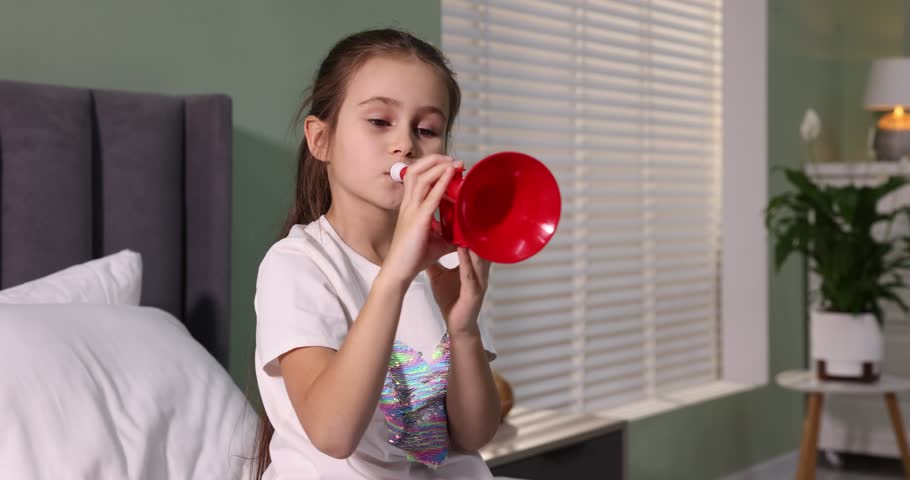Little girl playing toy trumpet on bed at home