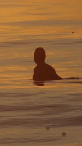 Surfer silhouette sitting in ocean sunset. lone surfer waits for incoming wave as warm golden light bathes.Vertical video
