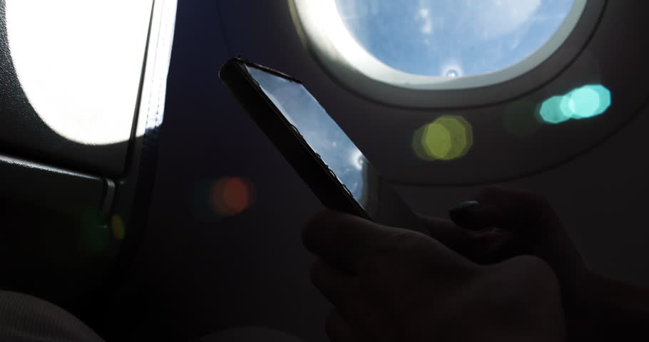 A close-up view shows a female hand holding a phone against an airplane window, as clouds and blue sky are visible outside. The moment captures a sense of travel and adventure.