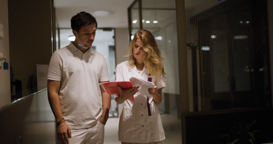A dentist and a nurse engage in conversation while reviewing patient files together in the quiet hallway of a contemporary reception area during their evening shift.