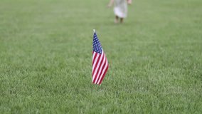 A small American flag stands upright in the middle of a wide green field as a child runs out of focus in the background - Powered by Shutterstock - Get 15% off with code: PIKWIZARD15