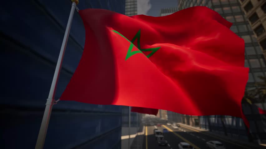 Morocco national flag waving along a modern city street under clear daylight, surrounded by buildings, palm trees and light traffic within an urban environment. The deep red textile, featuring the green five-pointed star known as the Seal of Solomon, commands attention against the azure sky. Located in North Africa, this Kingdom represents a blend of Arab and Berber cultures, often celebrated on Independence Day. As the banner undulates in the breeze, the emerald pentagram remains distinct, symbolizing the five pillars of Islam. Behind the ensign, the metropolis bustles with activity, possibly resembling the avenues of Casablanca or the administrative districts of Rabat. The fabric snaps crisply, showcasing the vibrant crimson hue that denotes the dynasty of the Alaouites. Nearby, tall structures of glass and concrete reflect the sun, while date palms sway gently, adding a natural touch to the developed zone. Vehicles move rhythmically along the boulevard, blurring slightly to emphasize the sharp focus on the national emblem. The standard maintains a position of pride atop the mast, reacting to every gust of air with fluid motion. Shadows cast by the staff fall upon the architecture, marking the passage of time. This footage captures the essence of the Maghreb region, highlighting a patriotic display in a commercial hub. The material exhibits a high-quality weave, visible as the sunlight catches the texture of the cloth. Visitors to the region, from the Atlas Mountains to the coast, recognize these colors instantly. The pendant serves as a constant reminder of sovereignty and heritage amidst the daily routine of the populace. The scene offers a neutral, observational perspective suitable for news broadcasting or travel documentaries focusing on the economy and state affairs of this historic land.