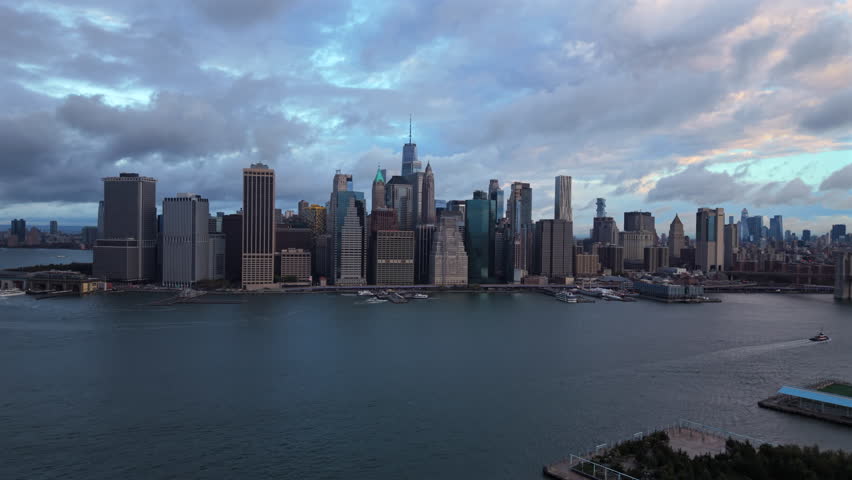 Drone aerial view of New York City, New York showing waterfront edges, tall skyline towers and soft cloudy daylight spreading across the river and urban scene.