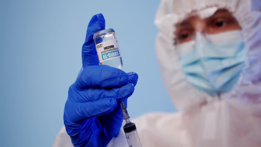 Blurred medical professional wearing PPE holds a syringe, capturing a healthcare setting, vaccination preparation, and medical safety procedures in a clinical environment.