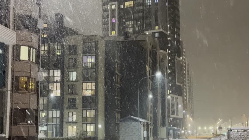 Heavy snow falls around modern residential buildings at night, illuminated by streetlights and window glow, creating a cold urban atmosphere with drifting flakes and muted winter tones.