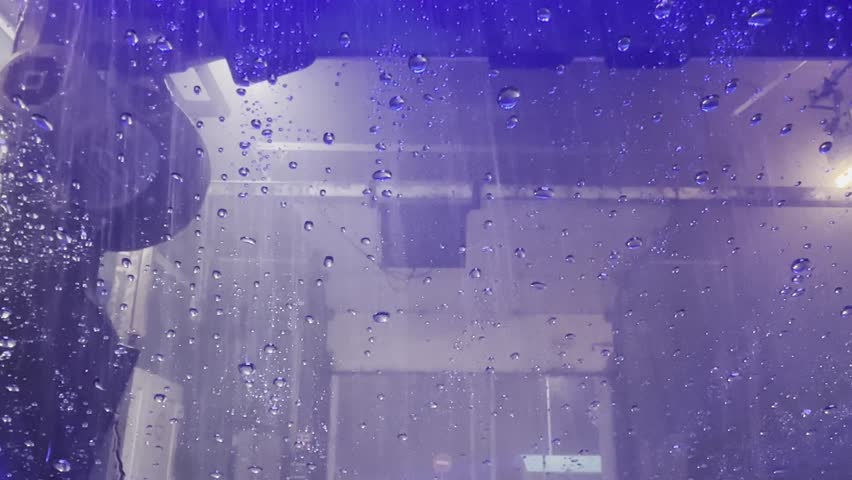 Water streams across the windshield during a car wash cycle, creating moving droplets and mist on the glass as the rinsing phase covers the surface in flowing patterns.