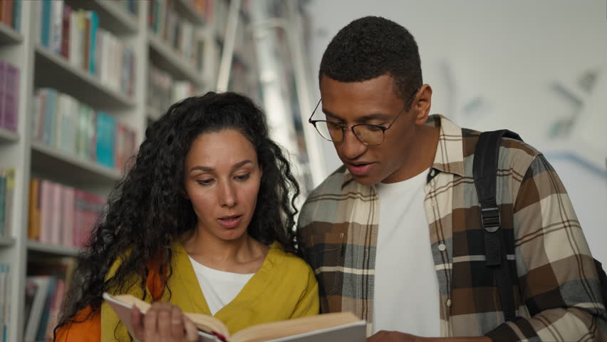Hispanic woman African American man in library reading book together education exam couple preparation learn study team discuss talk university textbook girl guy students friends bookworm conversation
