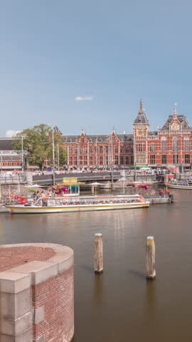 Timelapse of Amsterdam Central Railway Station front view with tour boats moored at the jetty, trams passing, and a canal in the foreground. Amsterdam, The Netherlands