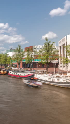 Boats floating and passing by near the National Opera music and theatre venue timelapse. Houseboats on the river in Amsterdam under a cloudy sky. Popular tourist destination in the Netherlands