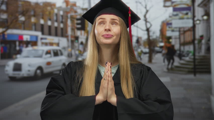 Woman in graduation cap and gown standing outdoors on city street with hands in prayer, surrounded by buildings and taxi, symbolizing hope and future aspirations.
