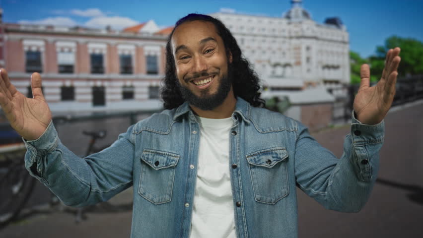 Man with beard smiling and open hands wearing denim jacket and white tee on a sunny street with parked bicycle; joyful welcome.