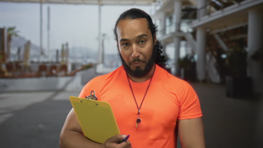 Man wearing orange shirt holding yellow clipboard while right hand makes ok sign in building; safety approval.