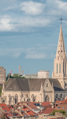 Aerial timelapse of Saint Servatius Church in Schaerbeek, Brussels, Belgium. Neo-Gothic spire surrounded by red-roofed houses under a blue cloudy sky. High-rise buildings and cranes in the background