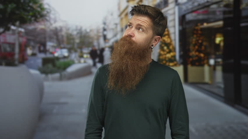 Man with long ginger beard looks up at sky on a busy city street with blurred background; curiosity.