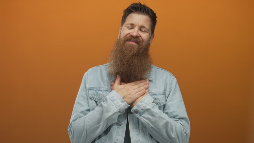 Man smiling with thick beard touching chest and closed eyes looking relaxed against orange studio wall; gratitude.