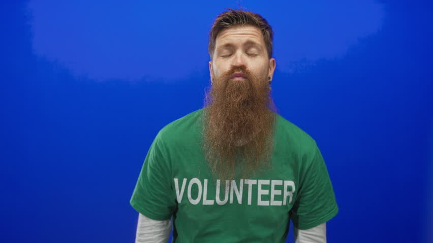 Young bearded volunteer man making finger heart gesture and blowing a kiss in studio; love affection.