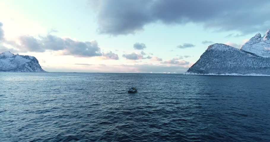 Cinematic aerial drone footage of a small fishing boat on Arctic sea with dramatic snowy mountains and soft pastel sky near Senja in Northern Norway, perfect for travel and adventure content.