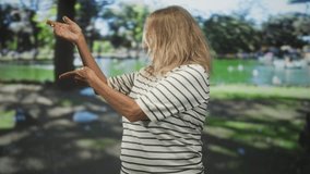 Senior blonde woman in striped shirt gestures with open palms and raised hands in a studio with projected park pond backdrop; serenity reflection. - Powered by Shutterstock - Get 15% off with code: PIKWIZARD15