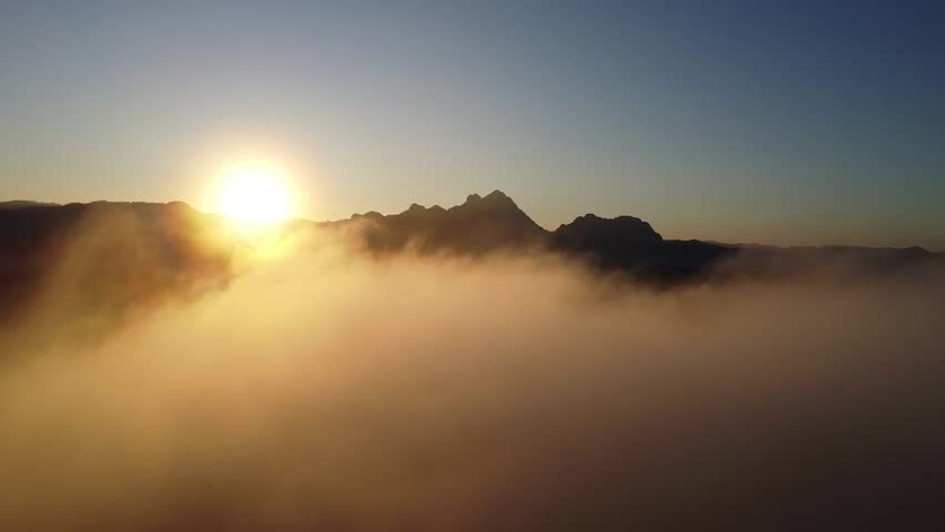 Aerial drone footage flying above soft clouds revealing a beautiful sunrise over mountain peaks with warm golden light.