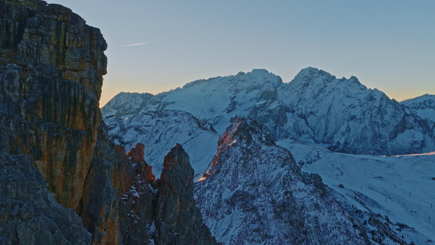 Cinematic aerial drone view of the snowy Dolomites in Val Gardena at sunrise. Sun rays create dramatic lens flares over rugged winter peaks and rocky ridges. A stunning panoramic mountain landscape id