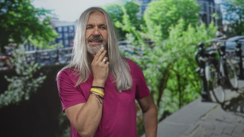Man with hand on chin on a street by a canal with bicycles, wearing rings and bracelets; contemplative.