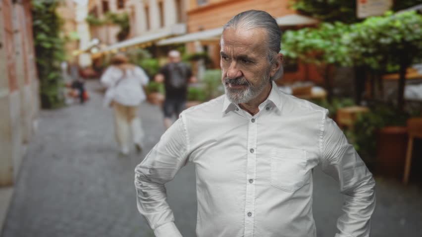 Man with hands on hips, hoary long hair tied back and gray beard, in white shirt on cobblestone street outside cafe; quiet contemplation.
