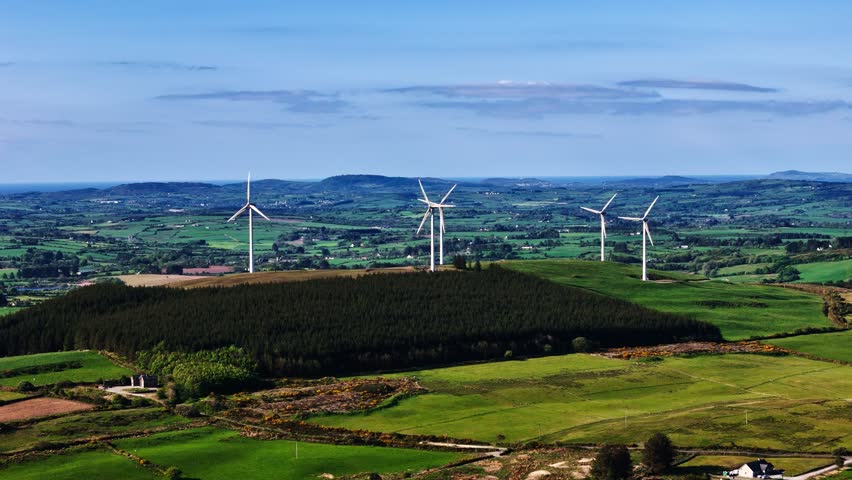Wind turbines are rotating slowly on a hilltop above a dark green forest. In the distance, green fields and mountains stretch to the horizon under a bright blue sky.