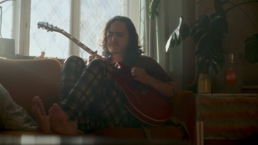 Young man with long hair sitting barefoot on couch, playing red electric guitar and enjoying music in cozy living room decorated with potted plant
