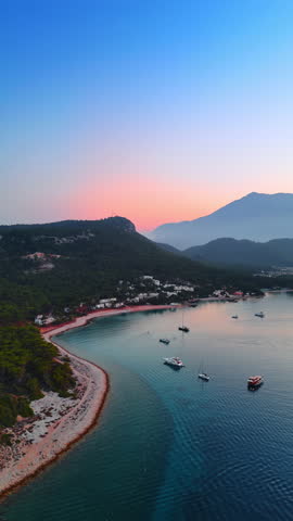 Coast of Turkey with a narrow line of sandy beach and green rocks. Some boats on the waterscape. Fethiye resort at sunset time. Vertical video.