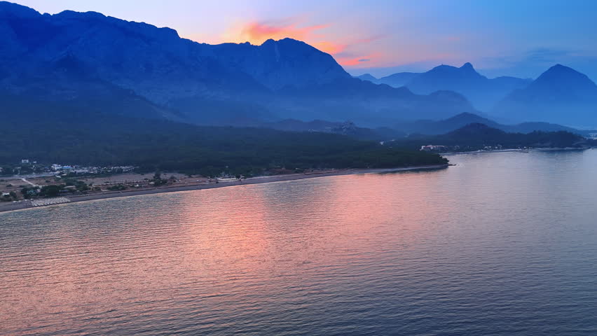 Last rays of setting sun color the waterscape pink from behind the mountain. Beautiful green shore surrounded by mighty rocks at dusk time.