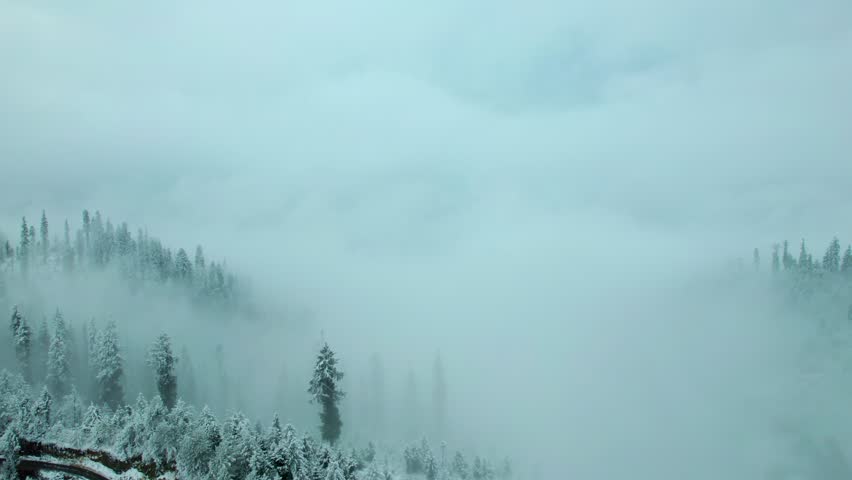 4K Aerial view of heavy storm clouds over snow covered mountain. Dramatic nature background. Fresh snow on trees and mountain during winter day.  Misty clouds rolling over the snow covered mountain.